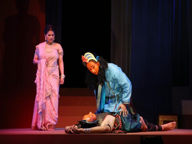 AKS TAMASHA 888 - bolebharat A theatrical scene: a woman in a pink sari stands on stage while a performer in blue with an orange mask crouches over a person lying on the floor.