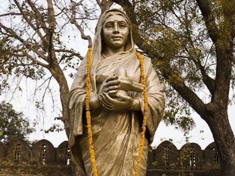 Statue of a veiled woman holding a bowl, draped with orange garlands, outdoors in a garden.