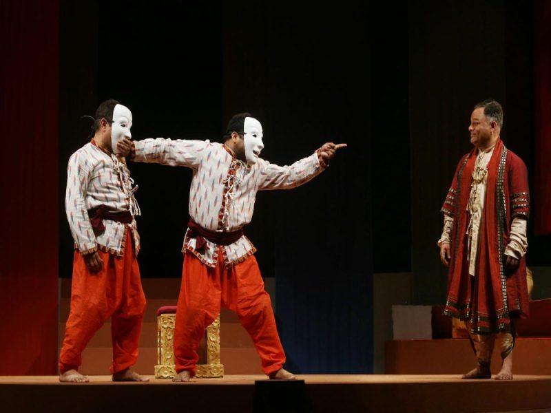 Aks Tamasha 2 - bolebharat Three performers on stage: two in white masks and matching patterned shirts with orange pants, one gesturing toward a third man in a red robe.