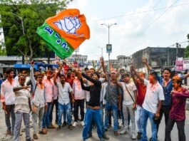 A crowd of men on a city street celebrate, with one man waving an orange-green flag featuring a white lotus symbol.