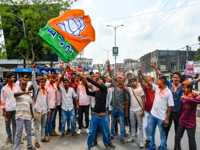 A crowd of men on a city street celebrate, with one man waving an orange-green flag featuring a white lotus symbol.