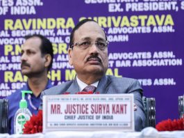 Man in a gray suit and glasses sits at a panel, nameplate reads 'Mr. Justice Surya Kant, Chief Justice of India'
