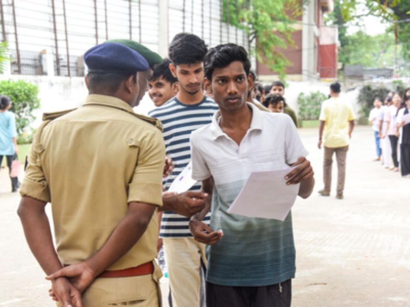 Nta ug exam cancelled - bolebharat Young man holding papers speaks to a uniformed officer outdoors while others wait nearby in a campus-like courtyard.