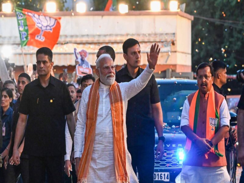 Indian political leader in a white kurta with an orange scarf waves to a cheering crowd at a rally, with security nearby and BJP banners in the background