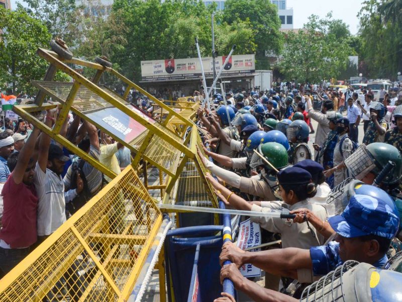 Crowd of protesters on a street confront police as yellow metal barricades are pushed forward by officers in riot gear with shields and batons.