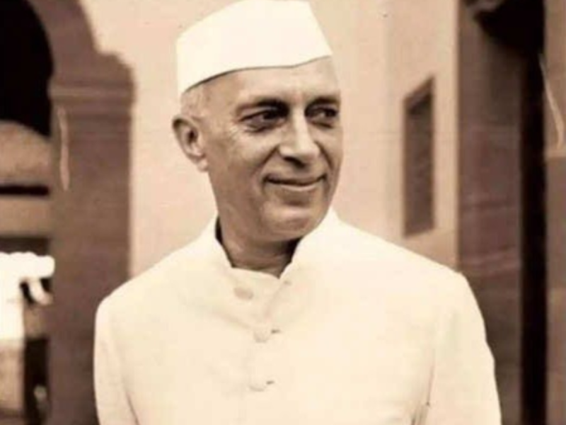 Older man in a white traditional Indian kurta and cap smiles while looking to the side indoors.