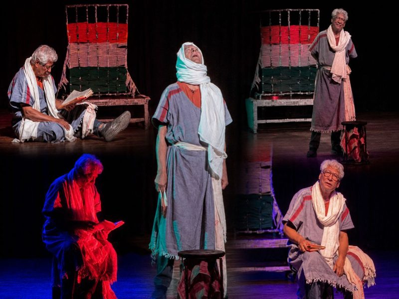 Stage theater scene with five performers in ancient-style robes; center figure stands wrapped in a white scarf, others sit or stand with props on a dimly lit set.
