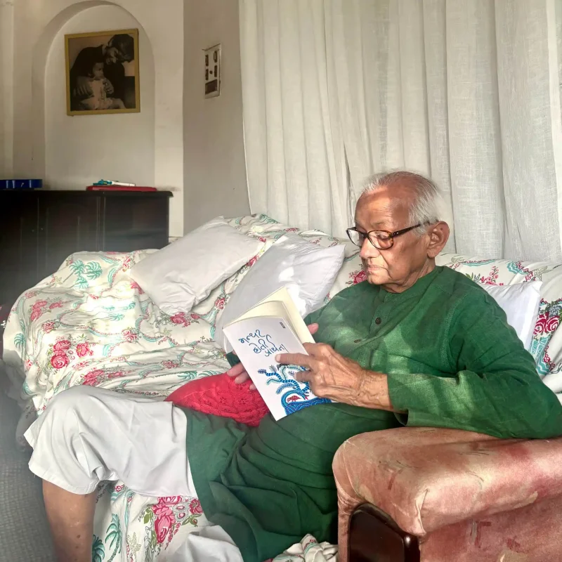 Two little things in the hands - bolebharat Older man with glasses in a green shirt sits on a bed, reading a card, with floral bedding and white curtains behind.
