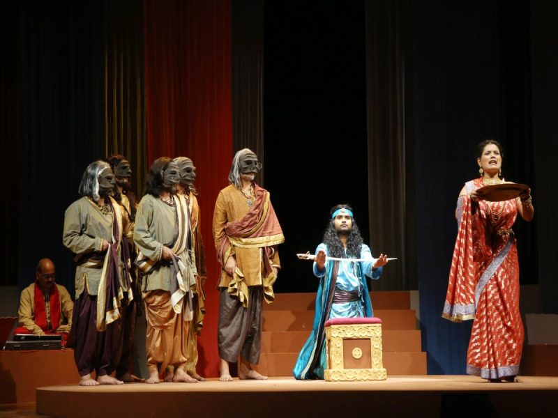 aks tamasha 3 - bolebharat Theater stage with masked actors in traditional Indian costumes; a blue-clad performer kneels center with a tray, while a woman in a red sari stands on the right holding a plate.