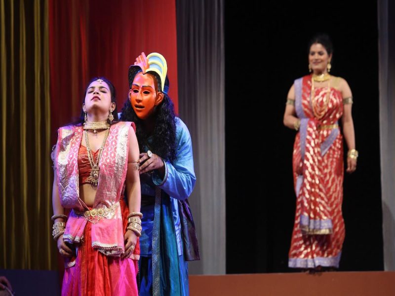 aks tamasha 7 - bolebharat On stage, a dancer in a pink traditional outfit stands with eyes closed as a blue-masked performer leans in behind her, while a woman in a red sari watches from the background on the right.