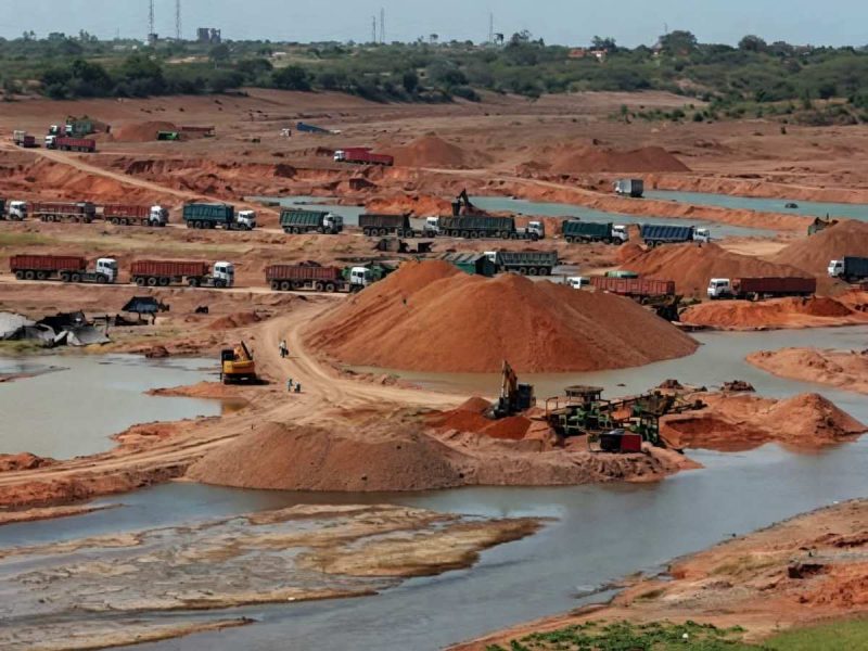 banda - bolebharat Active mining site with red soil, many dump trucks lined up and excavators working around piles of earth and water pools forming a quarry interior.