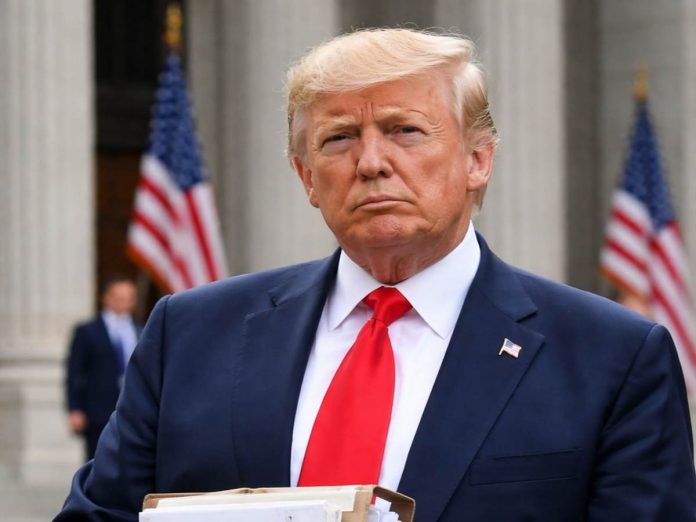 Man in a navy suit and red tie stands outdoors, holding a stack of papers, with American flags in the background and a serious expression.