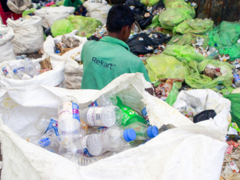 Person in a green shirt with back turned, sorting trash among large bags and heaps of plastic bottles and waste in a landfill setting.