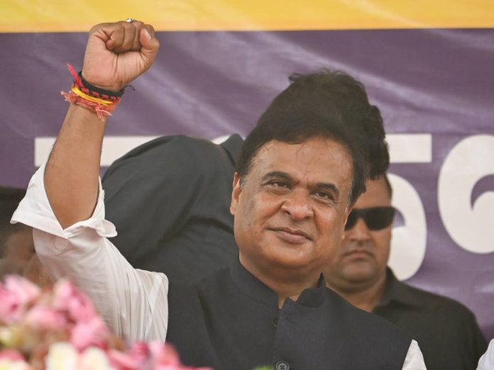 Man raises his clenched fist at a political rally, wearing a dark vest over a white shirt with bracelets on his wrist, purple banner behind him.