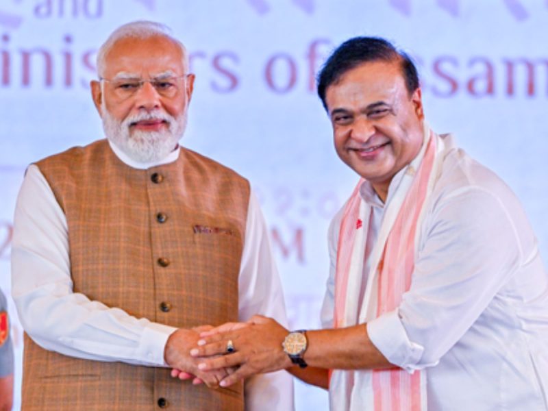 Two men on stage shake hands and smile at the camera, one wearing a brown check vest over a white shirt and the other in a white shirt with a light pink scarf behind them.