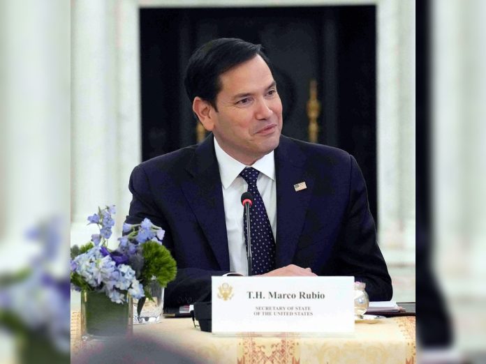 Marco Rubio in a navy suit sits at a table with a microphone; nameplate reads 'T.H. Marco Rubio' and a U.S. flag lapel pin is visible.