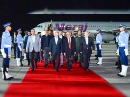 Dignitaries in suits walk down a red carpet at night after arriving by jet, flanked by uniformed guards on the tarmac.