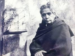 Older man wrapped in a dark blanket stands beside a rough wall; a birdcage is mounted to the wall.