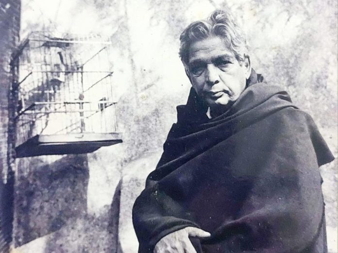 Older man wrapped in a dark blanket stands beside a rough wall; a birdcage is mounted to the wall.