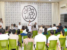 Audience seated in green chairs listening to a speaker at a podium in a conference room with a large patterned backdrop behind them.