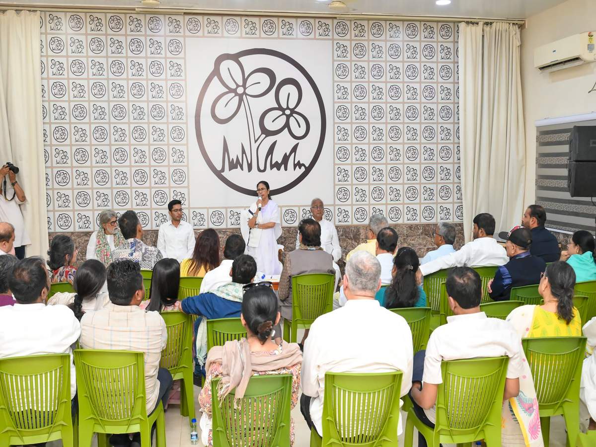 mamta banerjee tmc meeting Audience seated in green chairs listening to a speaker at a podium in a conference room with a large patterned backdrop behind them.