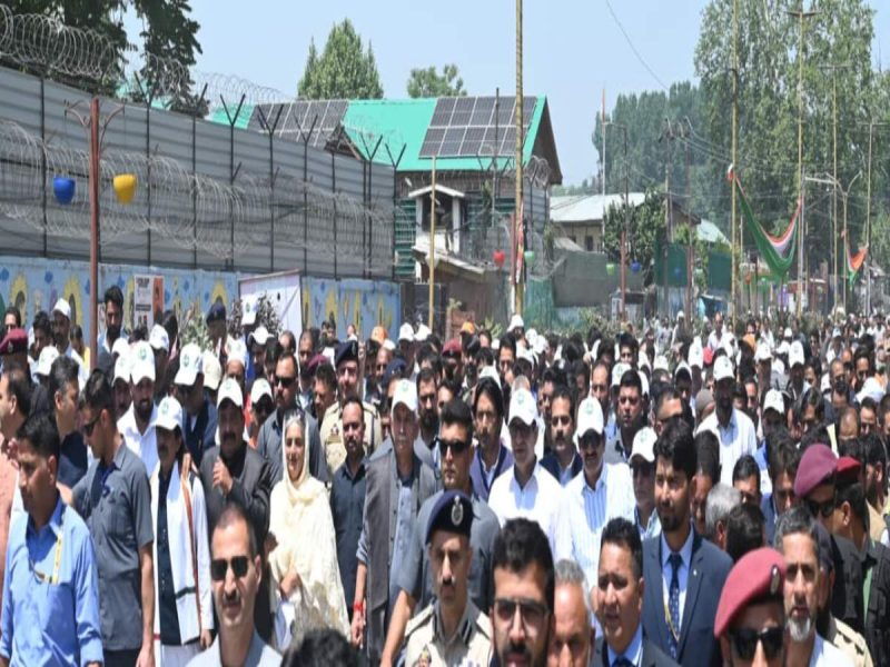 Large crowd of people marching along a street at a public event, with security personnel in front and barbed-wire fencing in the background.