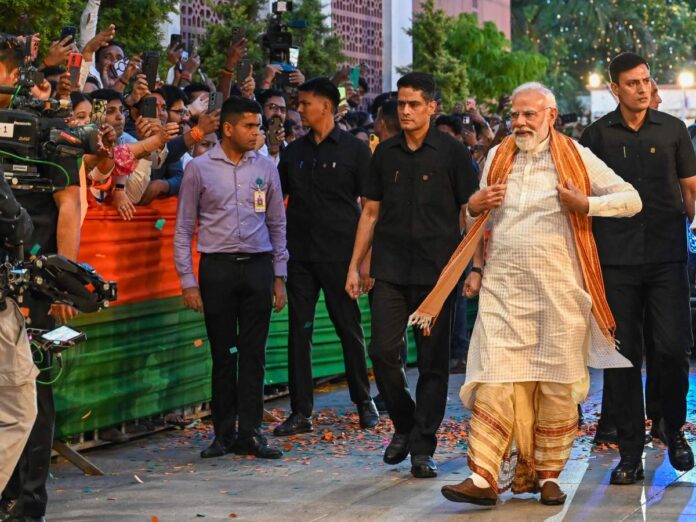 A dignitary in traditional attire walks past a cheering crowd and security guards while photographers snap photos nearby outdoors.