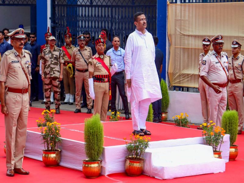 Man in a white kurta standing on a red-carpet podium during a formal ceremony, surrounded by uniformed police officers and guards.