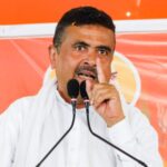 Man in a white shirt speaks at a podium with two microphones, raising his index finger during a rally or speech.