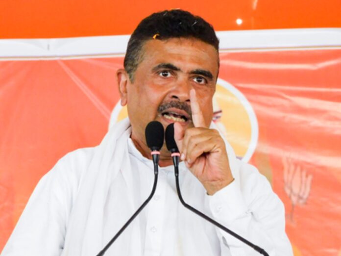 Man in a white shirt speaks at a podium with two microphones, raising his index finger during a rally or speech.