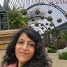 Close-up selfie of a woman with dark hair in a garden walkway with hanging moss balls and an arched metal trellis in the background.
