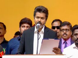 Man in a dark suit speaks into a microphone at a podium, holding a folder, with a bright orange backdrop and people behind him.