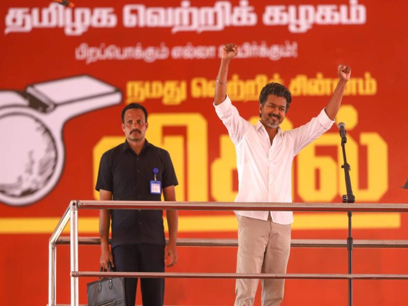 Man in a white shirt raises his arms on stage beside a microphone, with a security guard behind him against an orange Tamil backdrop.