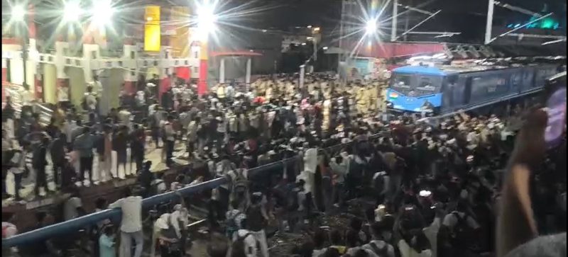 202606143824609 - bolebharat Crowded train platform at night with a blue passenger train pulling in and a dense crowd of people on the tracks and platform edges
