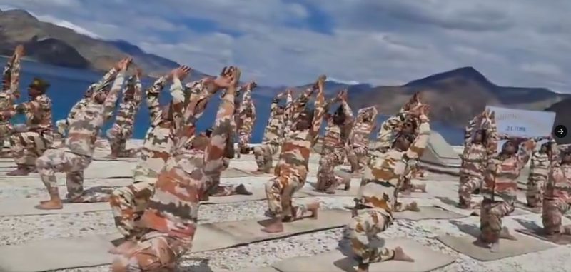 202606213832995 - bolebharat Group of soldiers in camouflage uniforms performing yoga or stretching poses on a rocky coastline with mountains and water in the background