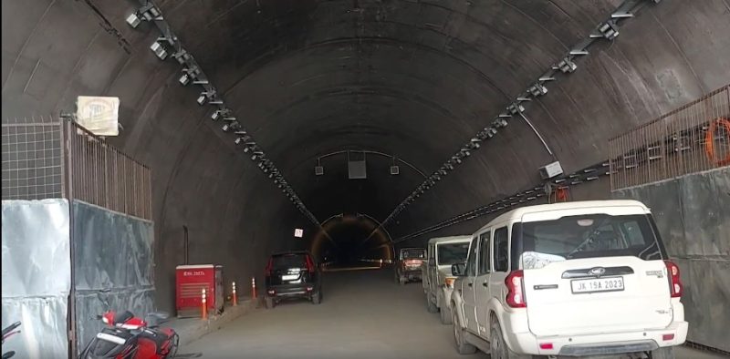 Vehicles parked inside a long, curved tunnel under construction with temporary lights and fencing along the walls.