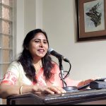 Woman in a peach and cream saree speaks into a microphone at a wooden podium.
