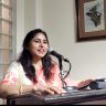 Woman in a peach and cream saree speaks into a microphone at a wooden podium.