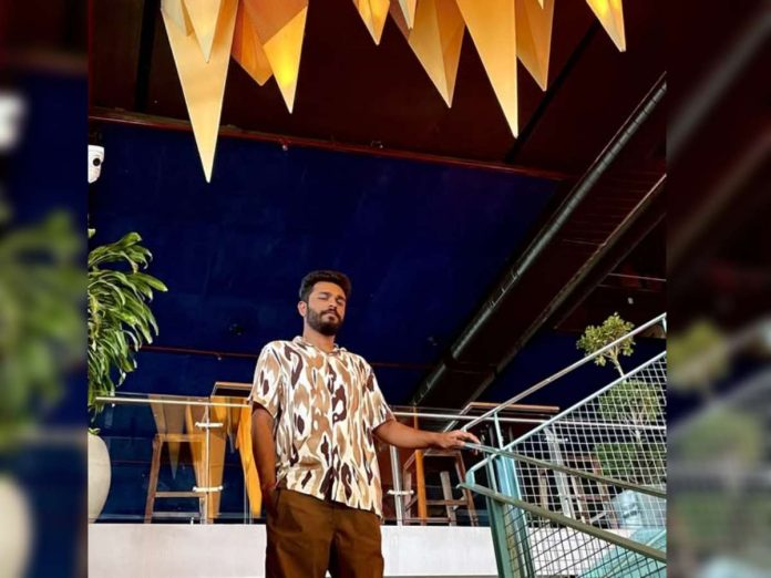 Man standing on a railing in a modern indoor space with a blue ceiling and yellow hanging decor, posing confidently.