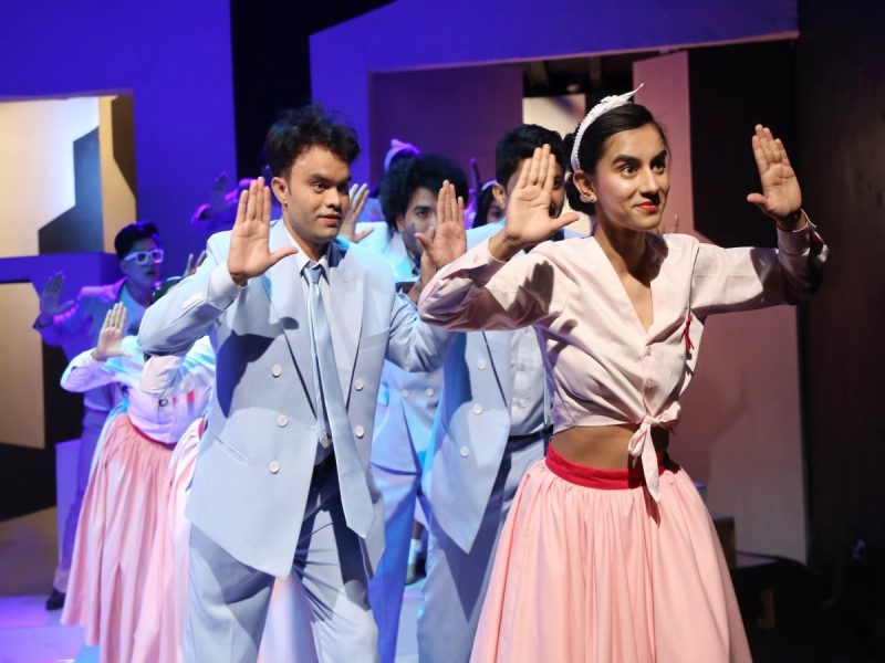 Dancers on stage performing a choreographed routine; man in a light blue suit and woman in a pink outfit raise their hands.