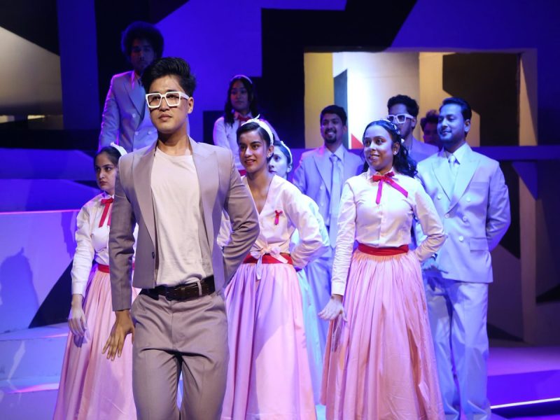 Lead male dancer in a beige suit and white glasses stands at the front as a group of dancers in white tops and pink skirts perform behind him on a blue-lit stage.