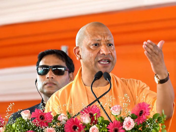 Speaker in orange robes addresses a crowd from a flower-decorated podium, gesturing with his right hand near two microphones.