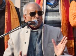 Elderly man wearing sunglasses speaks into a microphone at a microphone stand, gesturing with his hands in a crowd.