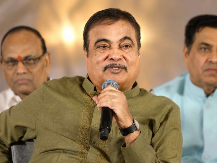 Man in an olive kurta speaks into a microphone at a panel or event, with two men seated behind him.