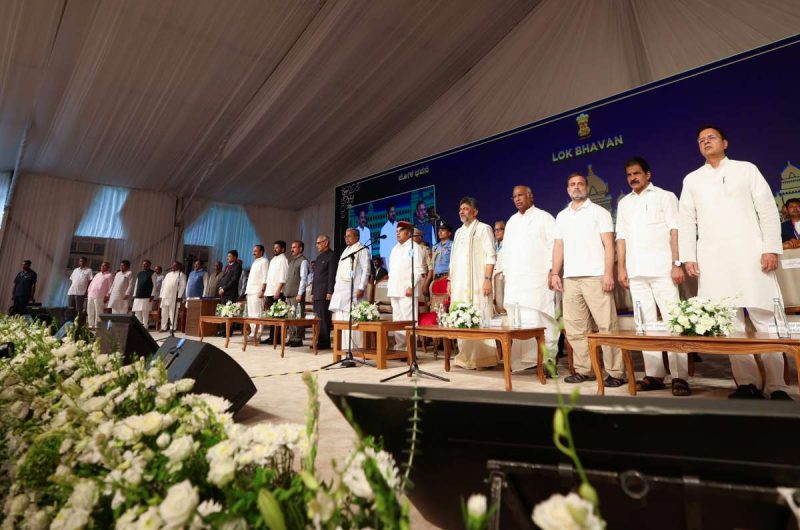 Bengaluru Swearing-in ceremony of Karnataka CM-designate DK Shivakumar - bolebharat A line of dignitaries stands on a stage under a white tent, facing the audience with microphones and floral arrangements in front.