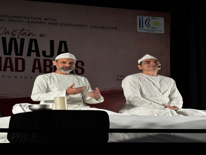Khwaja Ahmad Abbas 4 - bolebharat Two men in white traditional robes and caps sit on a stage with headset mics, speaking to an audience.