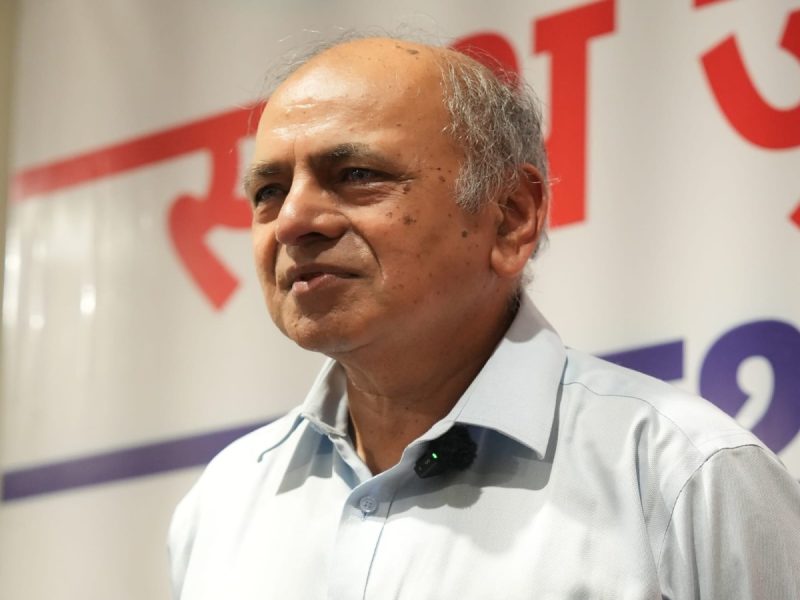 Close-up of a smiling man in a light blue shirt with a lavalier mic, speaking at an event with a red and purple banner behind him.
