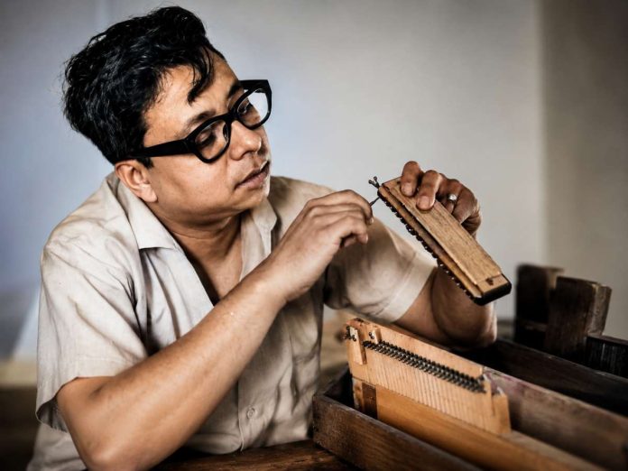 Man wearing glasses sits at a workbench, assembling a wooden stringed instrument.