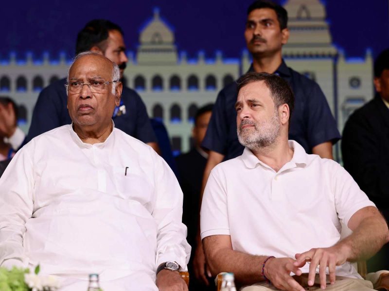 Rahul gandhi - bolebharat Two men in white shirts seated at an event, with a blue backdrop resembling arches behind them.