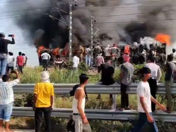 Crowd of bystanders watches a burning train on railway tracks, thick black smoke and orange flames rising behind a roadside guardrail.